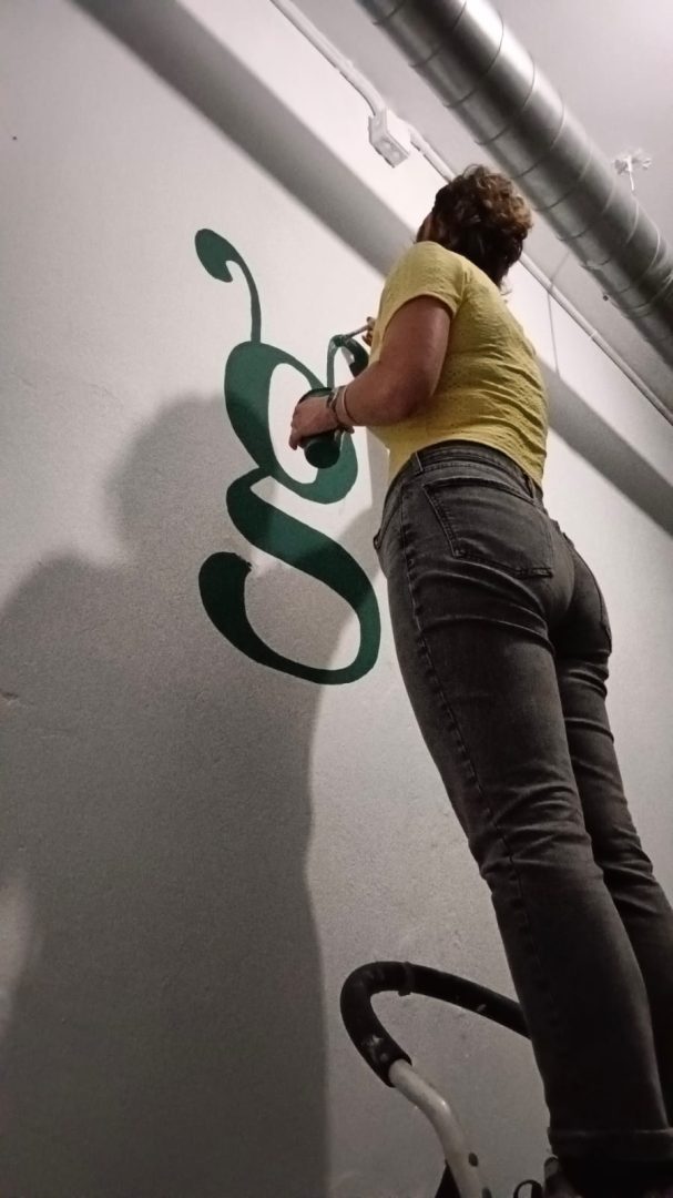 Persona pintando una letra grande en una pared, de espaldas, con pantalones ajustados.