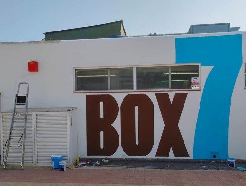 Muro blanco con texto grande: "BOX 7" en letras marrones y un número azul. Escalera de aluminio.